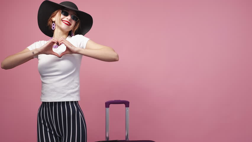 Sending love. Inspired woman. Travelling joy. Pretty happy lady in elegant summer look dancing with suitcase showing heart sign on pink background copy space. | Shutterstock HD Video #1098899379