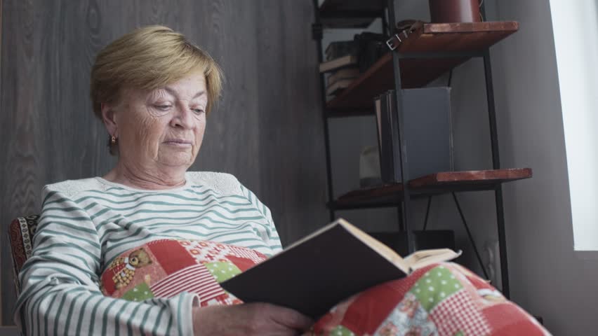 An elderly woman is reading in front of the window covered with a blanket. Old age and loneliness. | Shutterstock HD Video #1098904645
