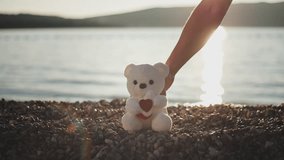 Man's hand puts white teddy bear with heart sign as gift on pebble beach in rays of setting sun on sunset. Valentine's day, Women's day, Mother's Day - Powered by Shutterstock - Get 15% off with code: PIKWIZARD15