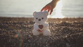 Man's hand puts white teddy bear with heart sign as gift on pebble beach in rays of setting sun on sunset. Valentine's day, Women's day, Mother's Day - Powered by Shutterstock - Get 15% off with code: PIKWIZARD15