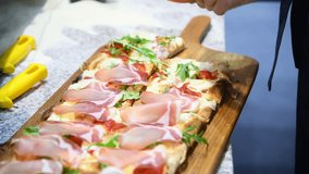 Close-up side view of cook putting sliced cured serrano ham on freshly cooked rectangular hot pizza on kitchen table. Real time video. Food industry theme.