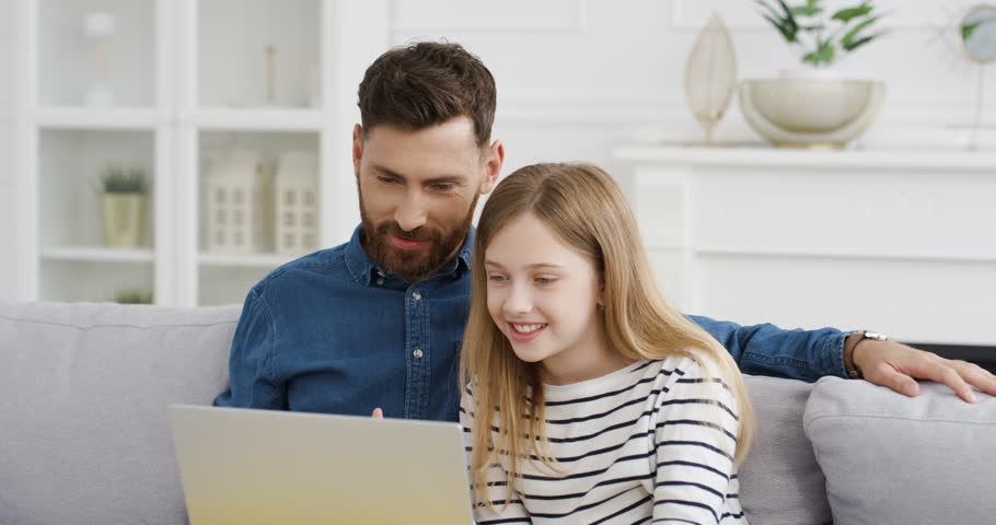 Caucasian father sitting on couch in living room and small cute daughter using videochat on laptop. Little girl with dad videochatting and giving thumbs up. Indoors. Child and daddy talking via webcam Royalty-Free Stock Footage #1098923713