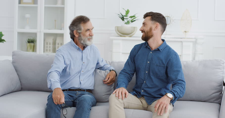 Caucasian males family generations. Happy cheerful senior father hugging his adult handsome son on sofa at home. Two men of different ages laughing and spending time together in living room. | Shutterstock HD Video #1098923737