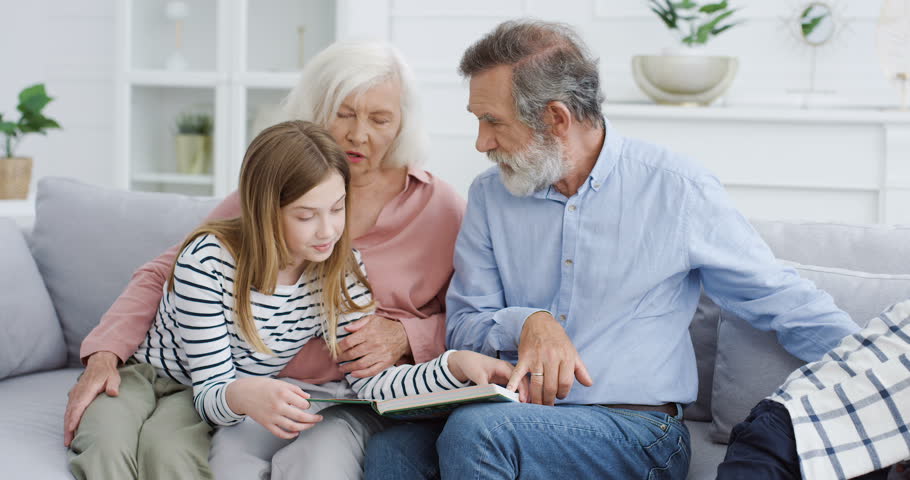 Caucasian old grandparents with grandchildren sitting on couch at home and reading book. Grandmother, grandfather, grandson and granddaughter spending time together and studying with textbook. Indoor.