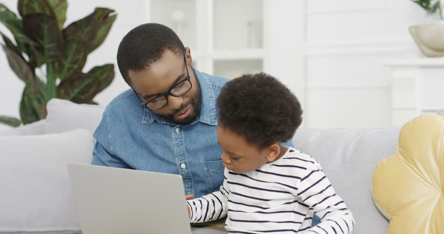 Young African American father sitting on couch in living room and teaching small cute daughter using laptop. Little girl typing on computer with her dad. Indoor.