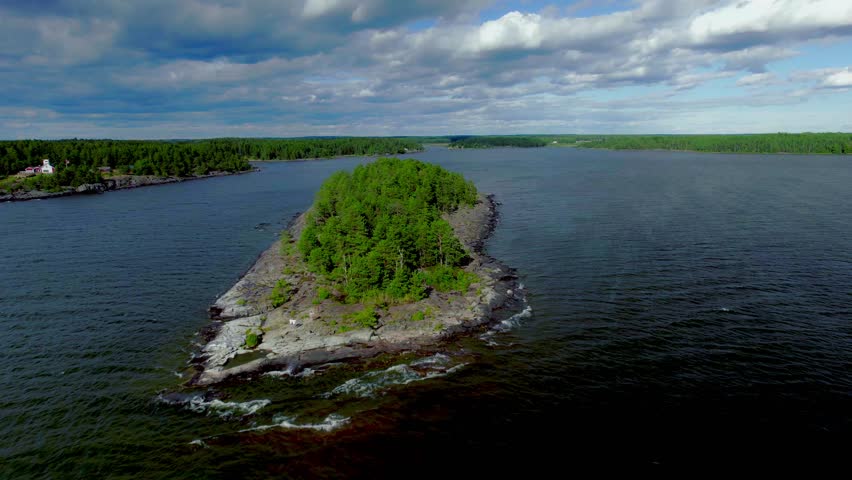 Low forward aerial over rocky island with pine trees. Islet in the middle of the lake with water waves, cloudy blue sky. Vänern lake in Värmland Sweden