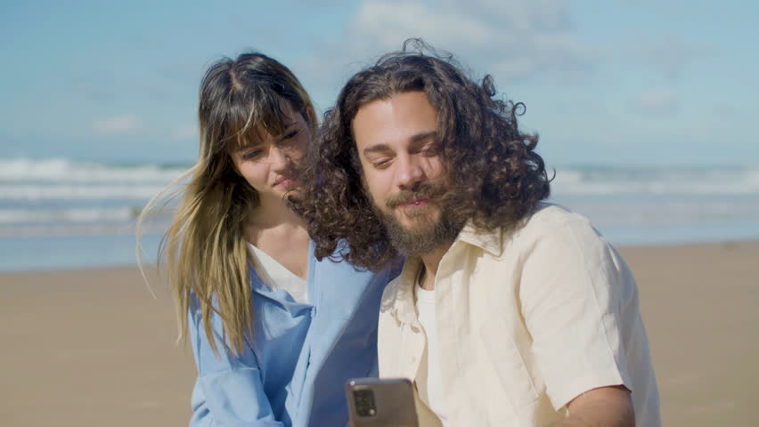 Beautiful couple taking selfie at seashore on sunny day. Bearded guy with long wavy hair and pretty blonde girl hugging while man holding phone and kissing girlfriend. Mobile technology, love concept