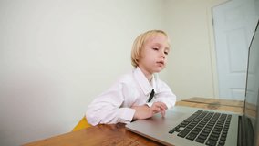 A preschooler boy dressed smart looks at the laptop screen and talks to a teacher repeating words and sounds. Online speech lesson at home for a 5 years old boy