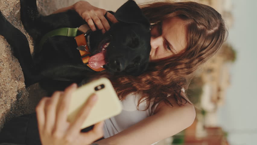 VERTICAL VIDEO: Clouse-up, cute girl with long wavy hair wearing white top takes selfie while sitting on the sand with black dog on modern building background. Girl hugging her pet | Shutterstock HD Video #1098933369