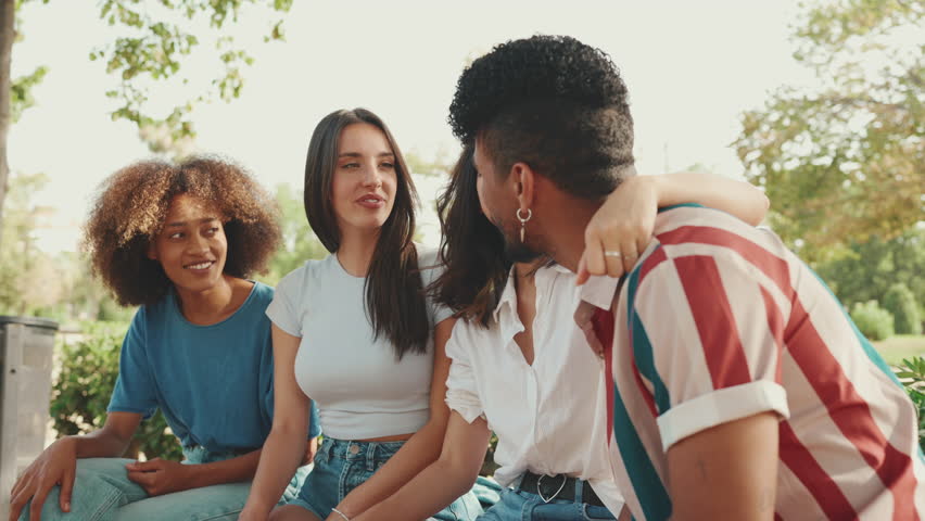 Happy multiethnic young people talking while sitting on park bench on summer day outdoors. Group of friends talking and laughing merrily in city park
