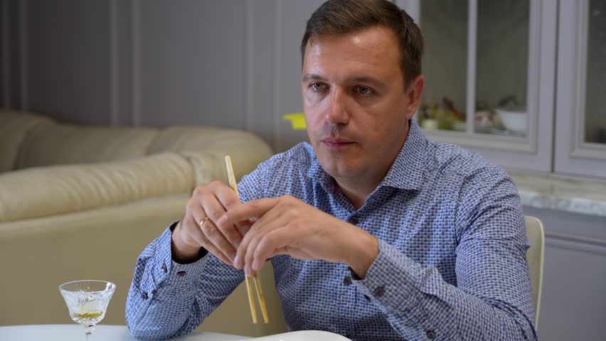 Millennial man in blue shirt communicates with his family sitting at dining table with chopsticks in his hands in dining room at home