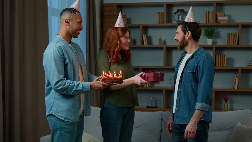 Ethnic couple congratulating adult friend wishing happy birthday presenting gift to bearded man in party cap blow out candles on cake makes wish celebrate anniversary with multiracial friends at home Royalty-Free Stock Footage #1098938517