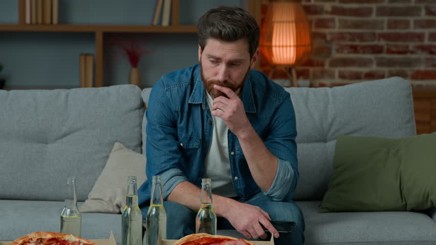 Portrait of upset adult man single caucasian male looking at pizza and beer sad unhappy guy lonely celebrate alone at home sit on couch no guests on birthday party feeling abandoned quarantine concept