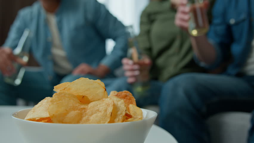 Close up plate with chips snacks on table young people men women friends sitting on couch indoors drinking beer together cheers clinking bottles tasting cold alcohol drinks celebrate Friday home party