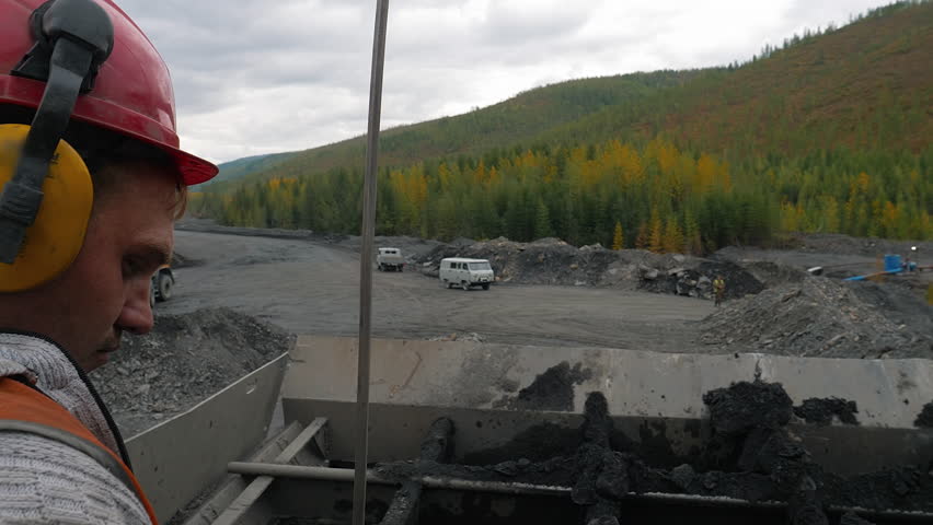 Mining Equipment Operator Watches Excavator Moving Mined Materials In Bucket. Mining Equipment Operator In Hard Hat And Safety Ear Muffs. Industrial Mining Equipment Observed By Operator