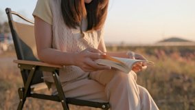 Video footage of Close up of woman is reading a book in meadow with mountain float on dam and sunset sky view.
