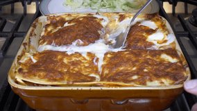 Closeup of a slice of homemade, freshly cooked, steaming hot lasagne being lifted from a ceramic baking dish on an oven hob, onto a plate of salad behind. Clip 3 of 3.