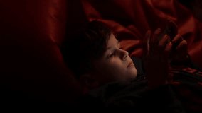 Little Caucasian boy 6 years old looks into a smartphone before going to bed in a dark room. Face of a child playing computer games, chatting with friends on social media lying on sofa in the evening.