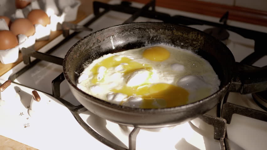 Female Hand is salting eggs in a hot skillet. Process of Cooking Scrambled Eggs in a Frying Pan in the Morning at Dawn. Bright sunlight. Sizzling oil, bubbles. Breakfast. Home kitchen. Egg set, tray. | Shutterstock HD Video #1098952843