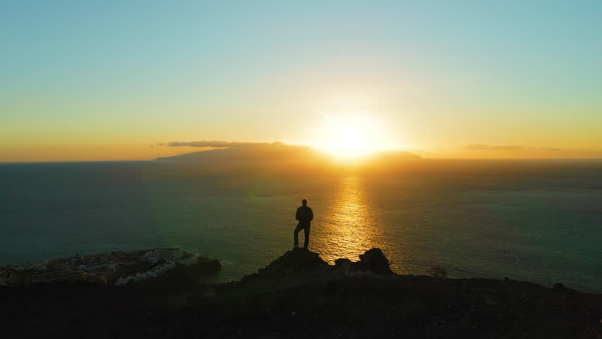Hiking man in mountains watching sunset and horizon over beautiful ocean water landscape. Male hiker silhouette against colorful orange sky. Sun glare, sunburst. Royalty-Free Stock Footage #1098955163