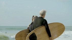 Elderly couple with surfboard walking along ocean shore. Back view of man and woman in wetsuit getting ready for surfing, spending time together. Family, retirement, extreme sport concept - Powered by Shutterstock - Get 15% off with code: PIKWIZARD15