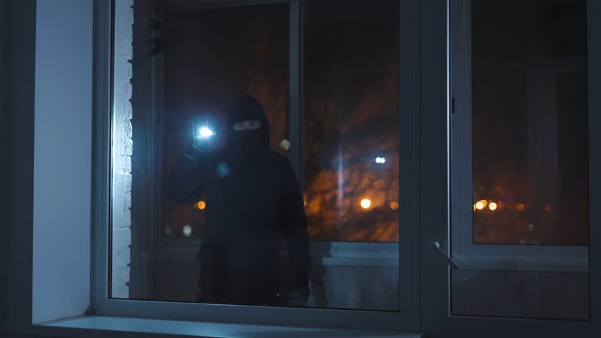 An unrecognizable burglar with a flashlight looks inside the house through the window. A male thief wants to break in and commit a theft during the night. Royalty-Free Stock Footage #1098966135