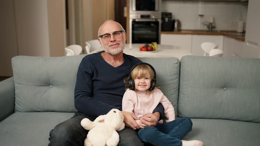 Portrait of a small girl in headphones with her grandpa while sitting on sofa