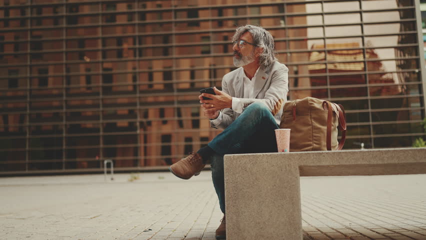 Mature businessman with beard in eyeglasses wearing gray jacket is using cell phone. Middle aged manager scrolling information on his smartphone while sitting outside the office