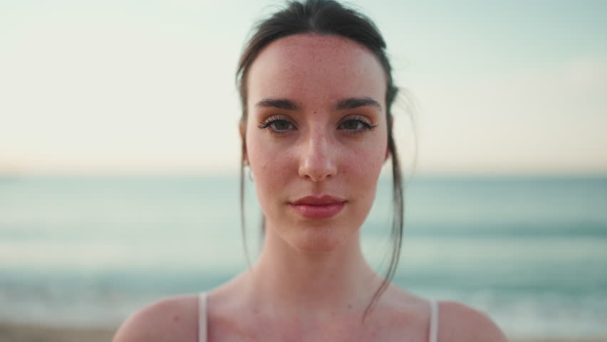 Close-up young athletic woman with long ponytail wearing beige sports top is standing eyes closed and relaxing on the sea background