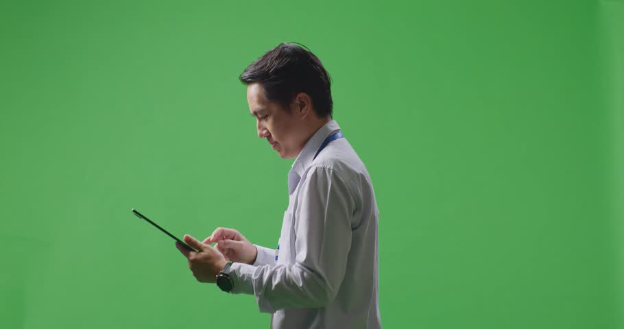 Side View Of Asian Business Man Using Tablet While Walking On Green Screen Background In The Studio
