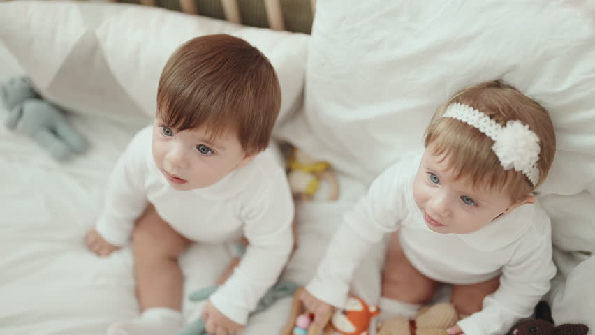 Two adorable babies holding toy sitting on cradle at bedroom