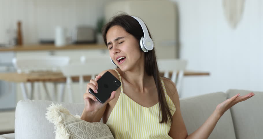 Beautiful young woman wear headphones rest on sofa, singing popular songs into smart phone using it like microphone, enjoy favourite track spend weekend at home. Carefree pastime, karaoke, fun, hobby Royalty-Free Stock Footage #1098976457