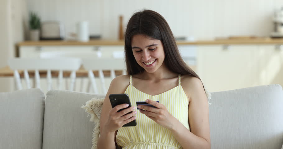 Attractive young woman enjoys e-shopping at home, sit on sofa holding credit card and smartphone, making order, buying clothes, goods, and gifts through e-commerce websites. Electronic commerce client | Shutterstock HD Video #1098976483