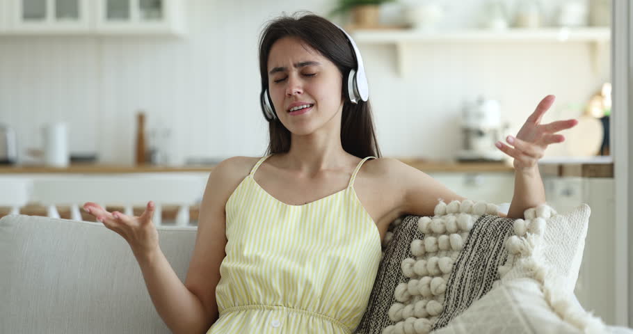 Attractive young 25s woman relaxing on couch in living room, sings song, listens to music through wireless headphones using on-line streaming services, enjoy hobby and carefree pastime alone at home Royalty-Free Stock Footage #1098976565