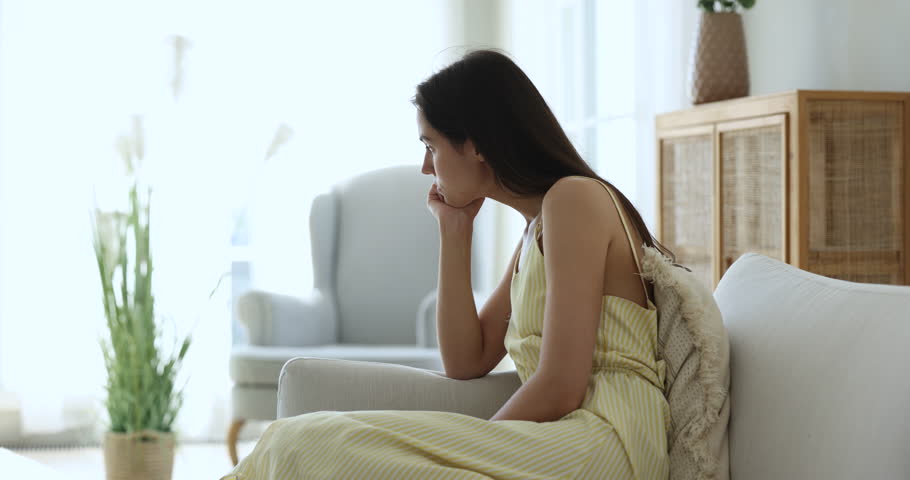 Young sad pensive female reflecting, suffers of loneliness, depression or boredom alone at home, feels fatigued resting seated on sofa staring into distance looks thoughtful, ponders, deep in thoughts Royalty-Free Stock Footage #1098976591