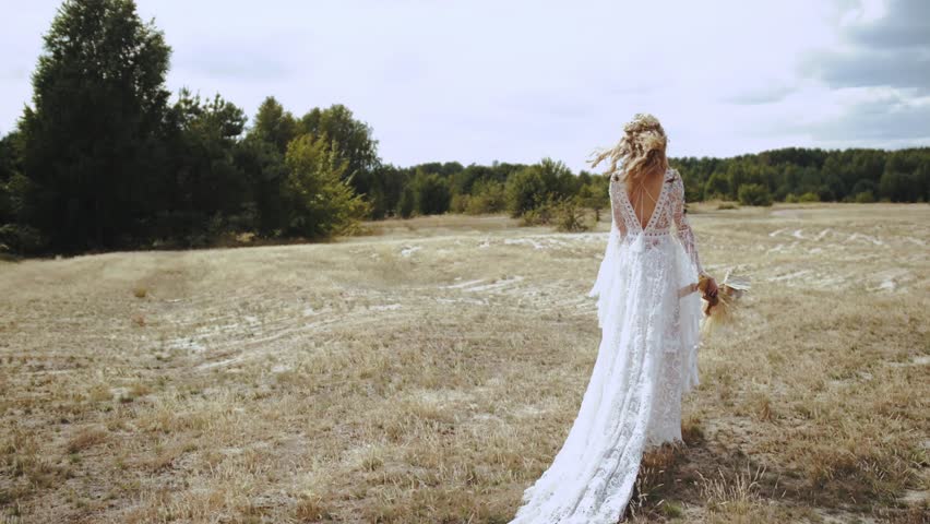 Bride in wedding boho dress walks through the field with a bouquet slow motion shot. The bride walks forward across the field and turns to the camera Royalty-Free Stock Footage #1098976685