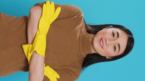 Vertical video: Smiling professional maid wearing protective rubber gloves standing with arm crossed in studio over blue background. Cheerful asian housekepper took great care in customer
