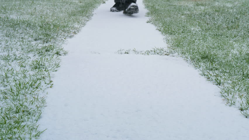 Closeup at ground level of a person’s shoes making the first footprints while slowly walking along a white, fresh snow covered path through grass, with gentle snowfall all around.