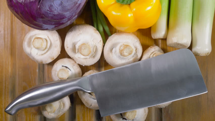 Overhead dolly of a steel cleaver at the start of a run of various fresh, raw vegetables on a worktop counter, ready to be cut or chopped, and a man’s hand pushing a cauliflower in place at the end. | Shutterstock HD Video #1098979507