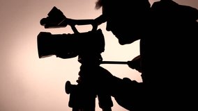 Closeup of a backlit man in silhouette leaning over to operate a video camera and perform a slow controlled pan shot, on a tripod in a studio setting, against a plain background.​
