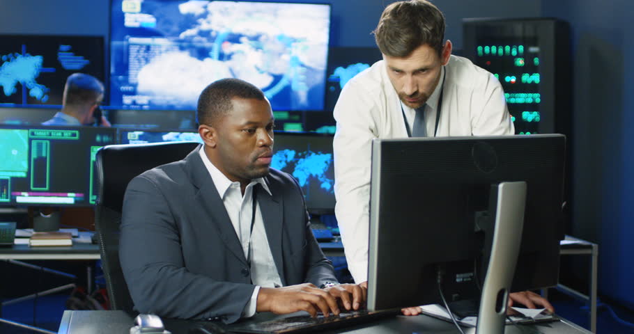 Two mixed-races males data security analytics talking and working at computer in military surveillance office. Multiethnic men, governmental officials discussing digital security in database center. Royalty-Free Stock Footage #1098981871