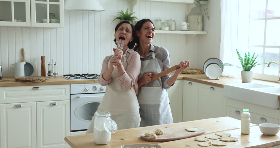 Vivacious happy mature woman and young adult daughter have fun, singing song into ladle and rolling pin, moving to favourite music enjoy cooking together in modern kitchen. Hobby, fun, home karaoke | Shutterstock HD Video #1098982673
