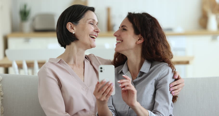 Adult Hispanic daughter and older 50s mother rest on sofa, holding smartphone, watch photos and videos, recollects, enjoy memories spend leisure together at home using gadget. Modern tech, e-commerce