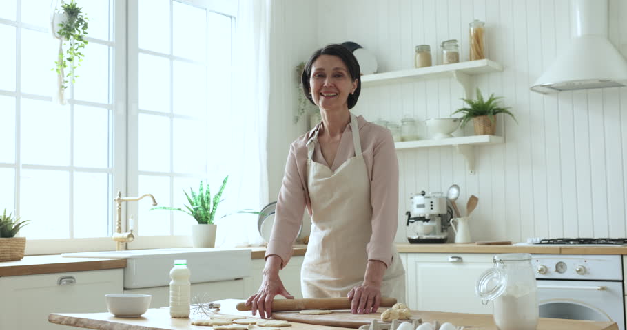 Positive retired mature brunette woman wear apron cooking alone standing in kitchen, prepare dough for homemade pastry smile staring at camera enjoy cookery process at modern home. Chores, housework | Shutterstock HD Video #1098982703