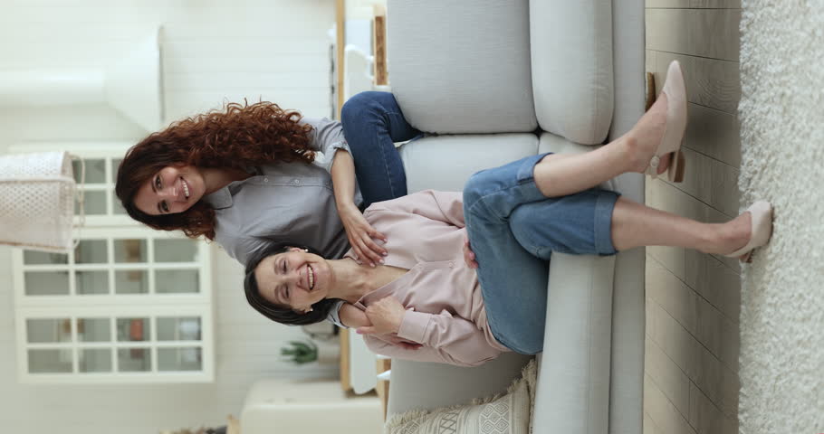 Two beautiful older and younger women, senior 55s mother shooting, looking at camera, posing together with young Hispanic attractive grown up 35s daughter sit on sofa looking at cam. Family portrait | Shutterstock HD Video #1098982717