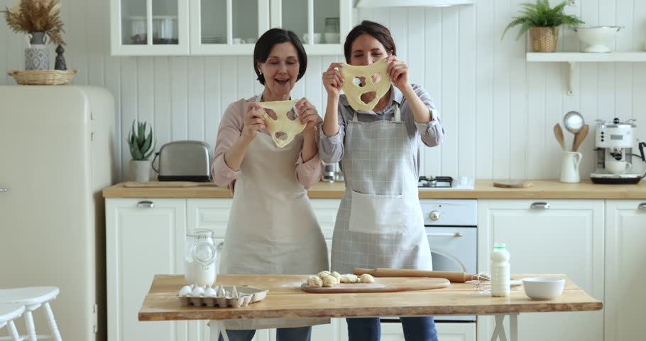 Grownup daughter her 55s mom in aprons cook together in domestic kitchen hold dough near faces like masks prepare homemade Halloween cookies joking, feel happy, laugh, have fun at home. Family pastime Royalty-Free Stock Footage #1098982725