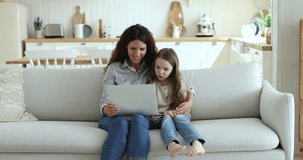 Young Hispanic woman and little cute daughter resting on sofa with laptop, watching educational vlog or listening favourite on-line music spend leisure together on internet. Fun, modern technologies