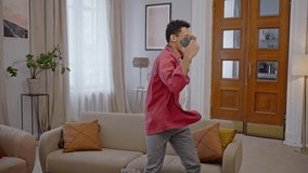 Slow motion. Young multi-ethnic man dancer, choreographer, performing modern freestyle dancing movements with a phone in hand, rejoicing, expressing his personality by circling around himself at home - Powered by Shutterstock - Get 15% off with code: PIKWIZARD15