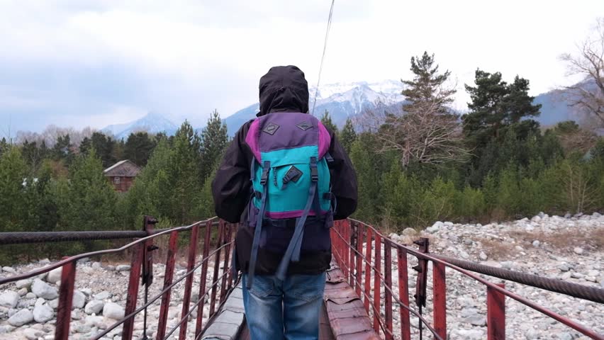 tourist with a backpack in an anorak with a hood walks over an old suspension bridge over a rocky mountain river towards the mountains. cloudy day. slow motion. back view. concept of travel.