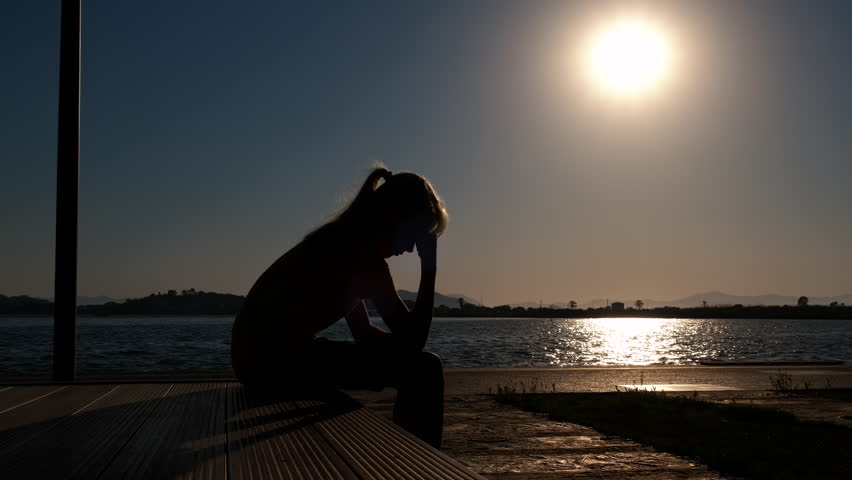 Girl lonely feeling silhouette by sea. A view of thoughtful young girl silhouette sitting by the sea in last sun rays. A concept of depression at nightfall.